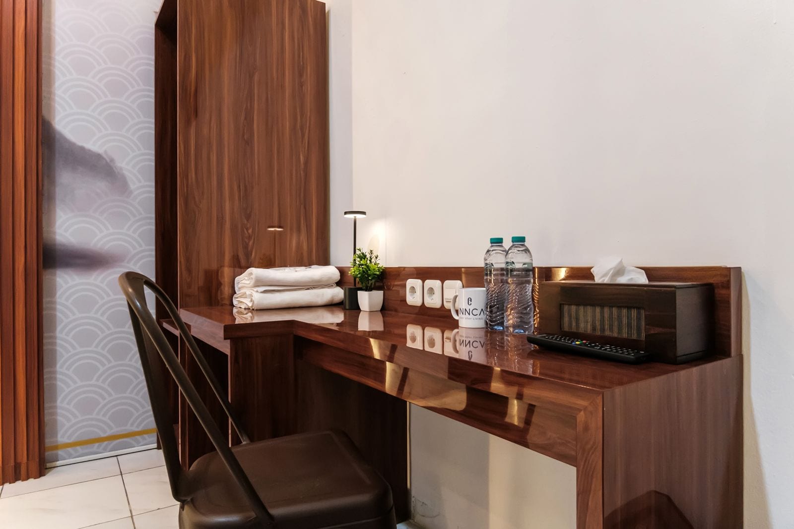 A hotel room with a wooden desk, a chair, a remote control, a radio, and a small potted plant.