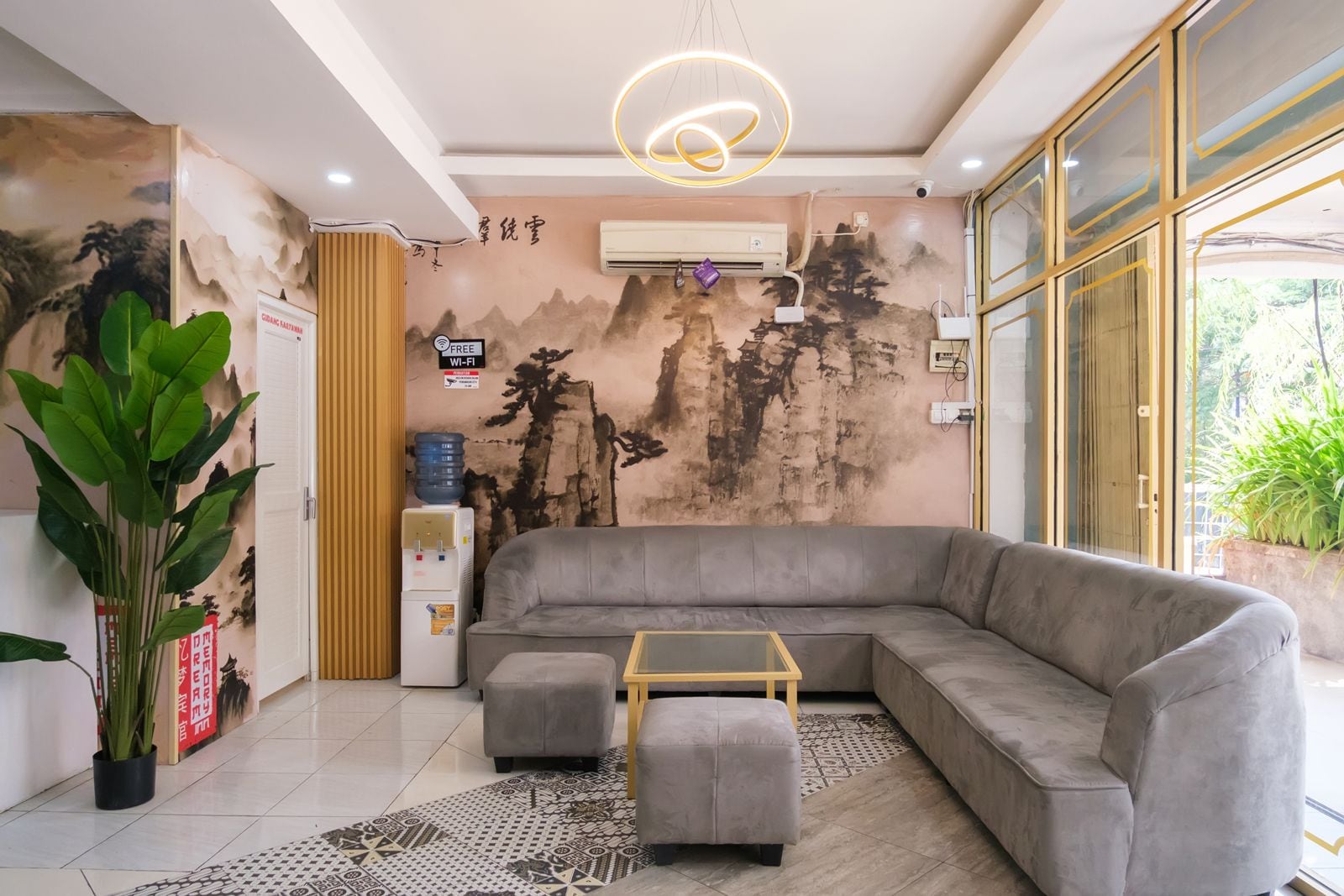A modern living room with a grey sectional sofa, a small ottoman, a glass coffee table, a water dispenser, and an air conditioning unit.