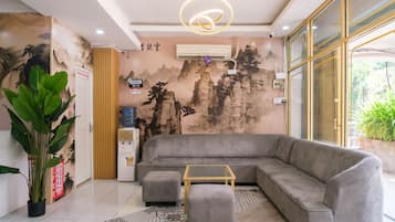 A modern living room with a grey sectional sofa, a small ottoman, a glass coffee table, a water dispenser, and an air conditioning unit.