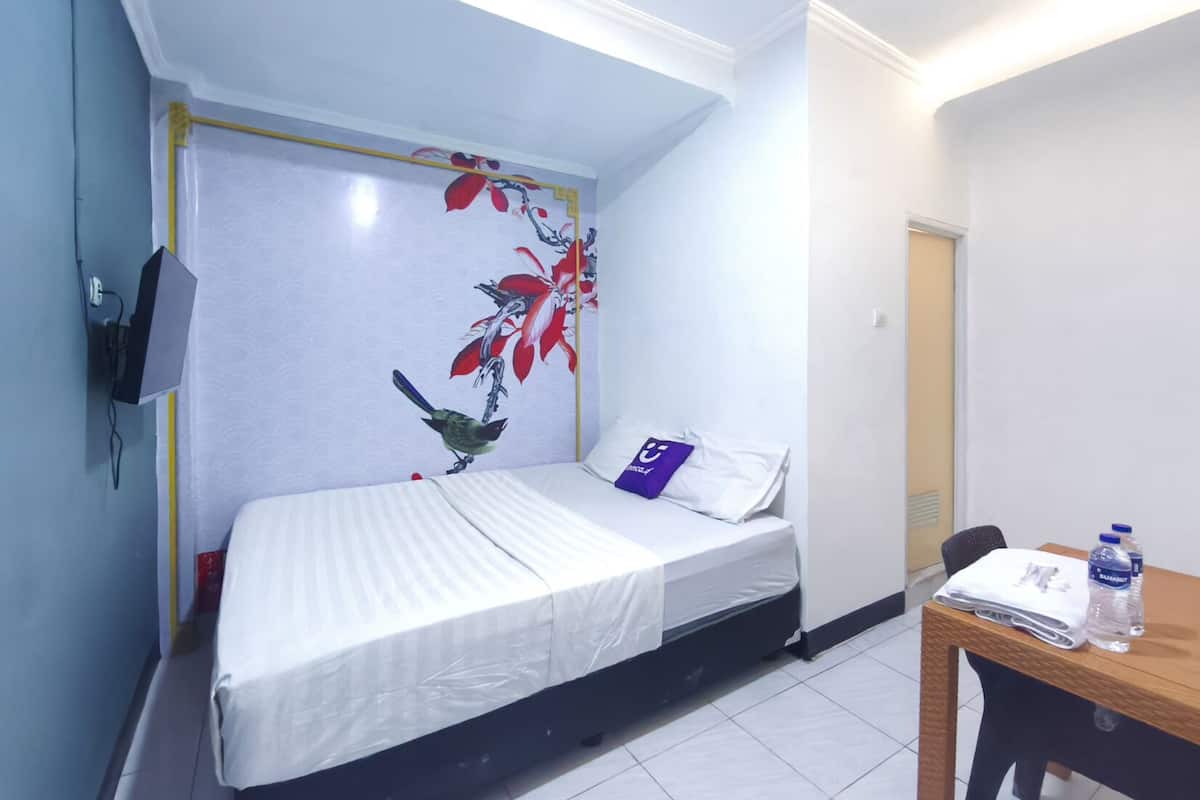 A small, neatly made bed with a white mattress and a single pillow, a small table with a water bottle, and a wall with a floral mural.