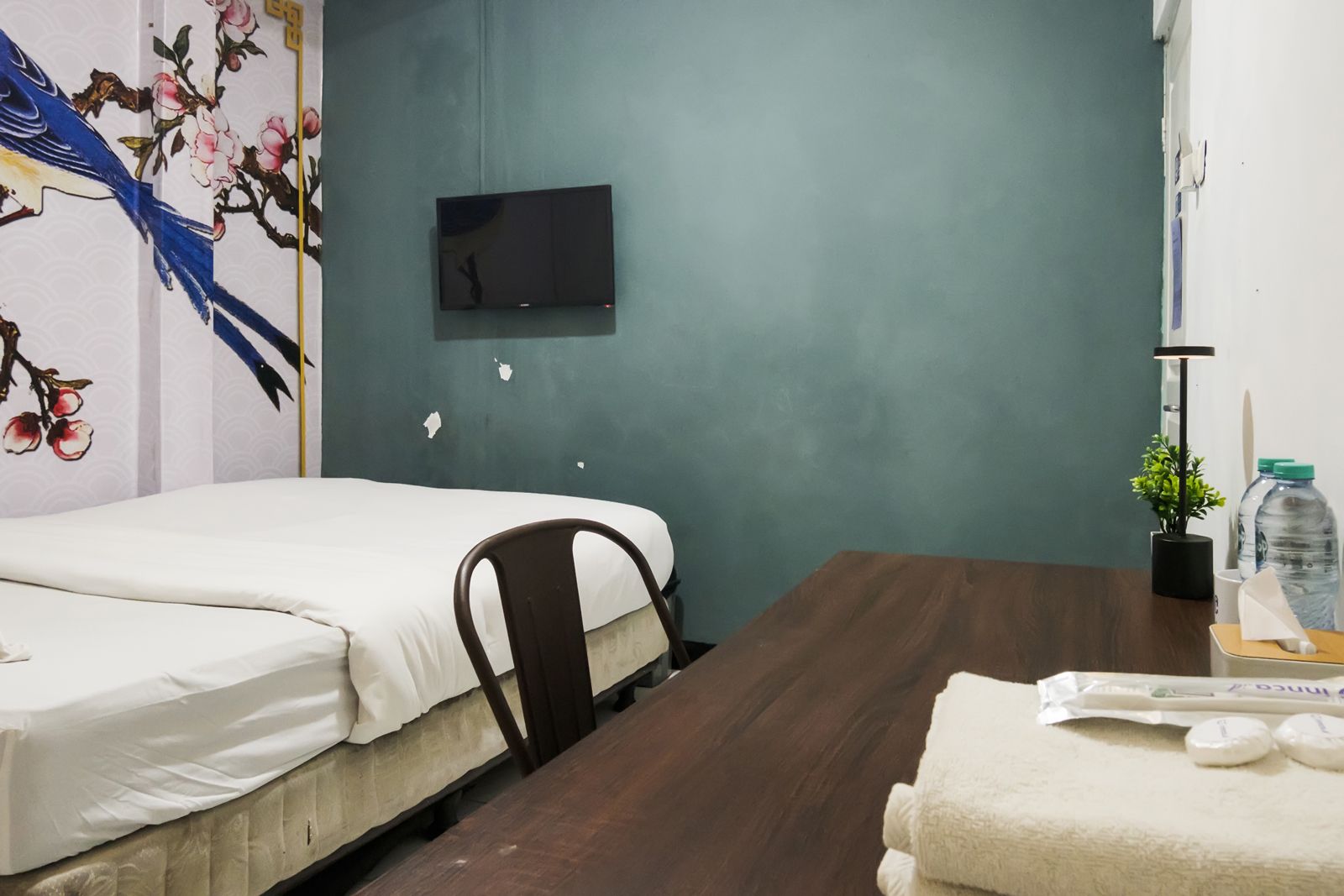 A hotel room with a bed, a chair, a table, a TV, and a wall with a floral painting.