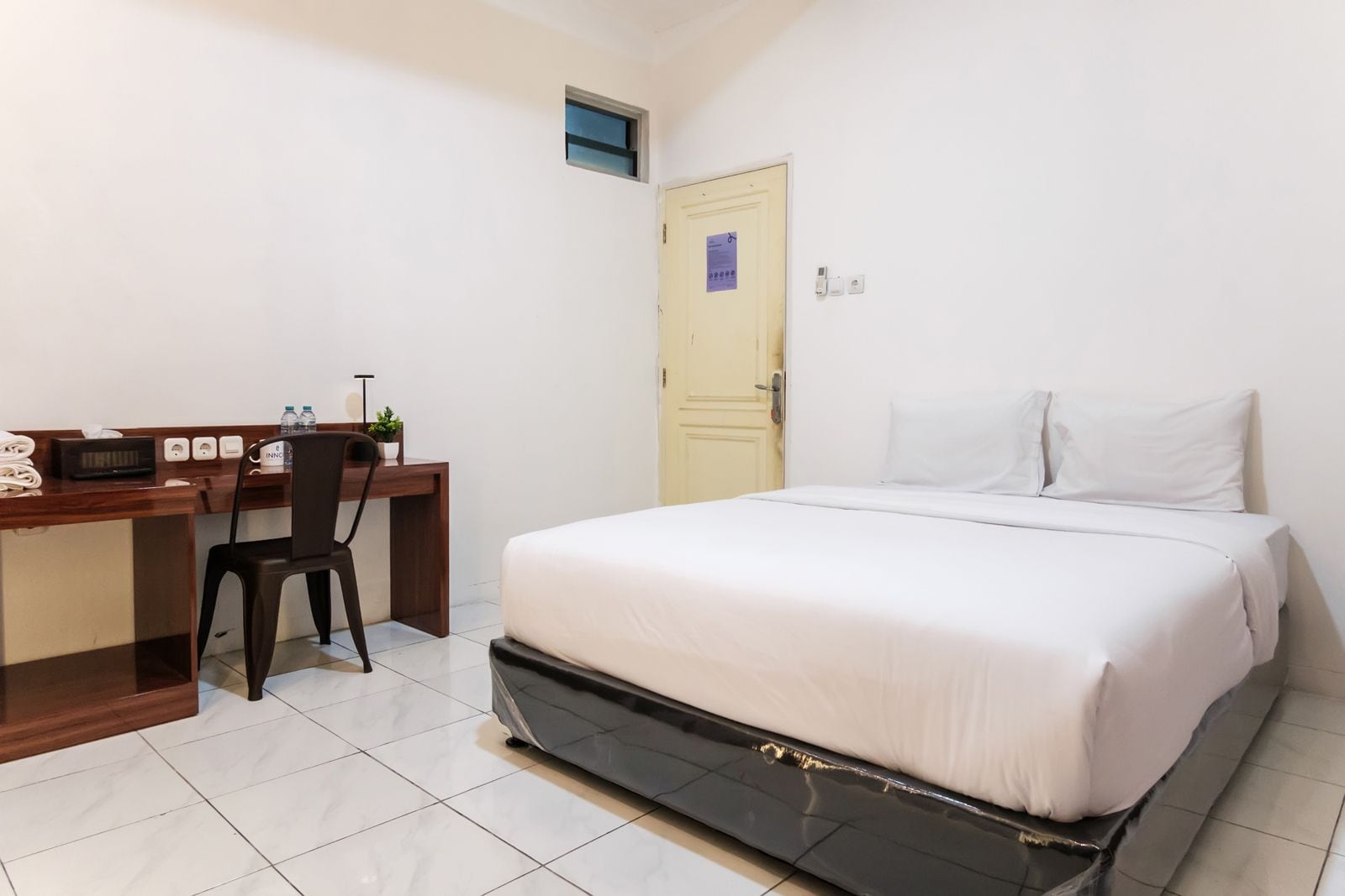 A hotel room with a bed, a desk with a chair, a small window, and a door.