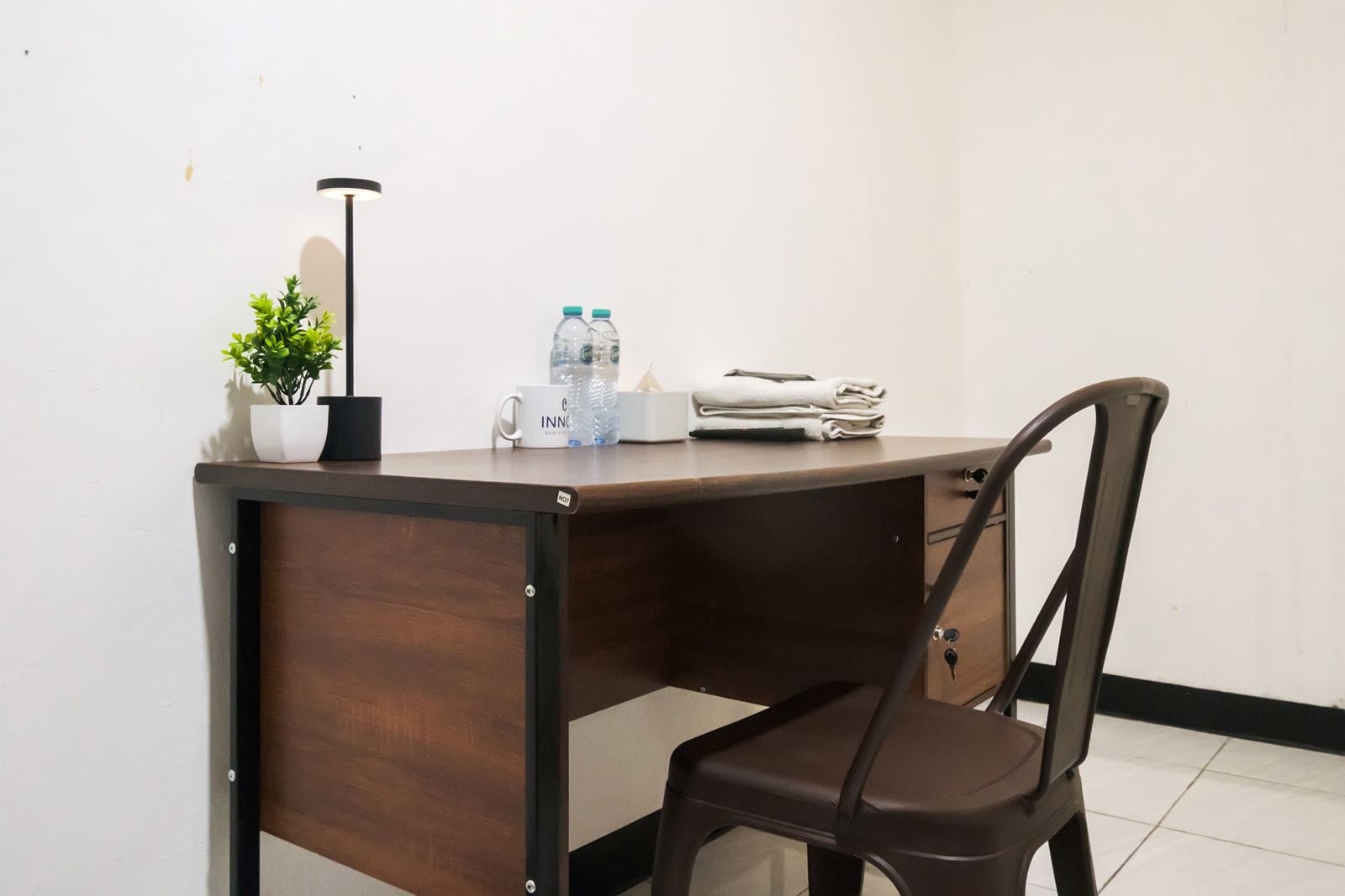 A small, dark wooden desk with a chair, a potted plant, a water bottle, a mug, and a folded towel.