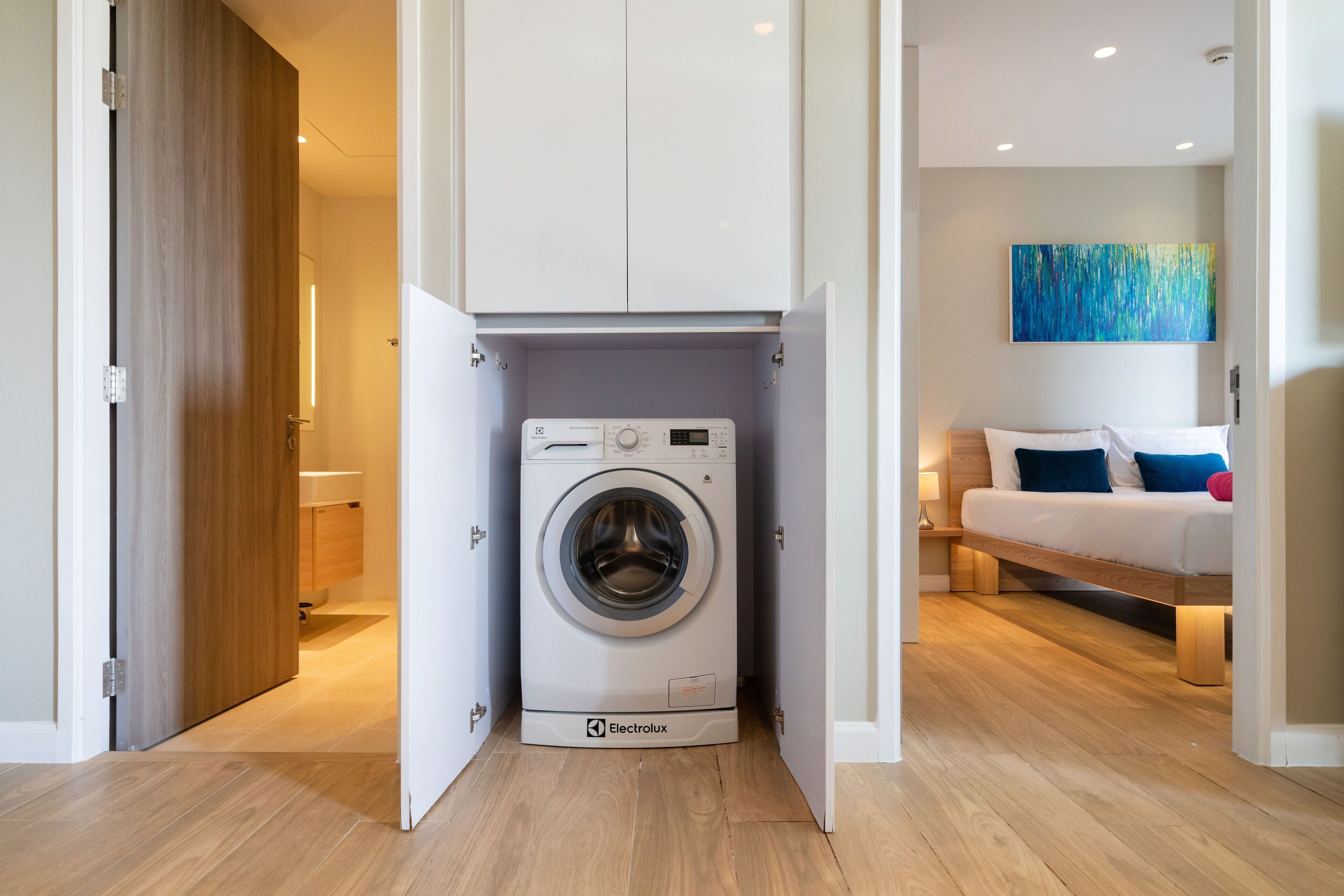 A compact, modern laundry room with a front-loading washing machine, a bed with a wooden frame and a blue pillow, and a painting on the wall.