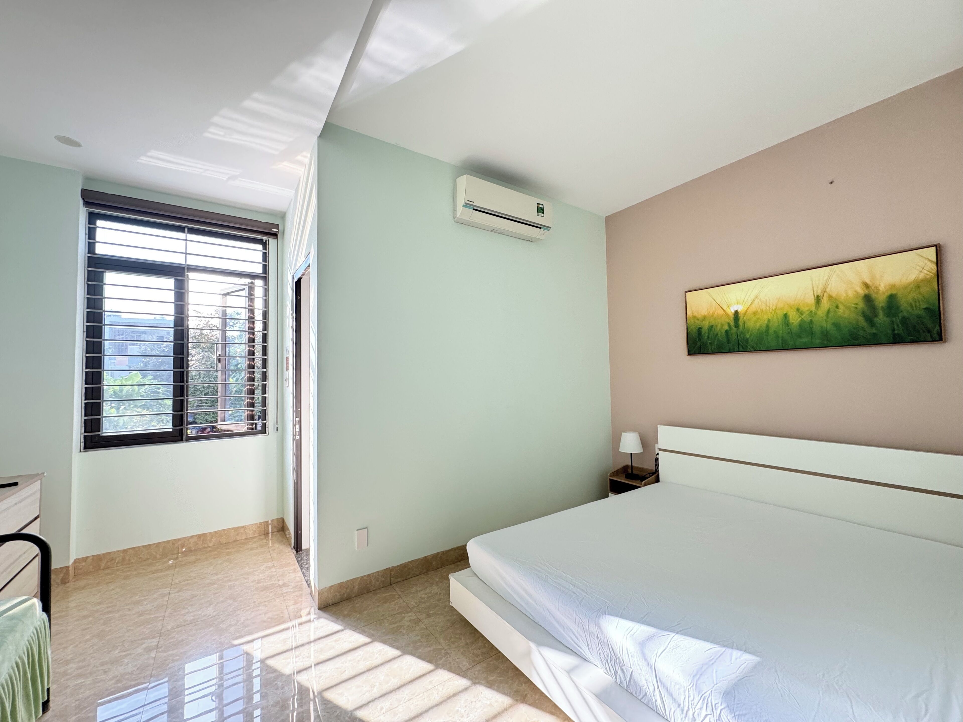 A bedroom with a bed, a window with blinds, an air conditioning unit, and a painting on the wall.