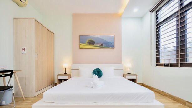 A neatly made bed with white linens and a teal pillow, a wooden wardrobe, a framed landscape picture on the wall, and a window with blinds. - Anh House (Da Nang)