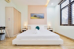 A neatly made bed with white linens and a teal pillow, a wooden wardrobe, a framed landscape picture on the wall, and a window with blinds. - Anh House (Da Nang)