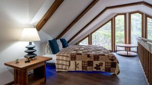 Ein Schlafzimmer mit schrägen Wänden, einem Bett mit einer Steppdecke, einem Nachttisch mit einer Lampe und Blick auf Bäume durch große Fenster.