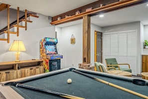 A pool table with a cue stick, a colorful arcade machine, and a green chair in a room with wooden beams and a white door. - Tranquil Lake Harmony Spa Chalet, Hot Tub, Game Rm (Lake Harmony)