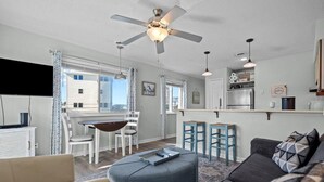 Ein modernes Wohnzimmer mit Sofa, Hocker, Esstisch, Stühlen und einer Küchenzeile mit Kühlschrank und Barhocker.
