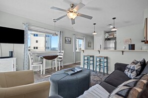 Ein modernes Wohnzimmer mit Sofa, Hocker, Esstisch, Stühlen und einer Küchenzeile mit Kühlschrank und Barhocker.