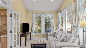 Ein Wohnzimmer mit Sofa, Hocker, Fernseher und einem Fenster mit Vorhängen.