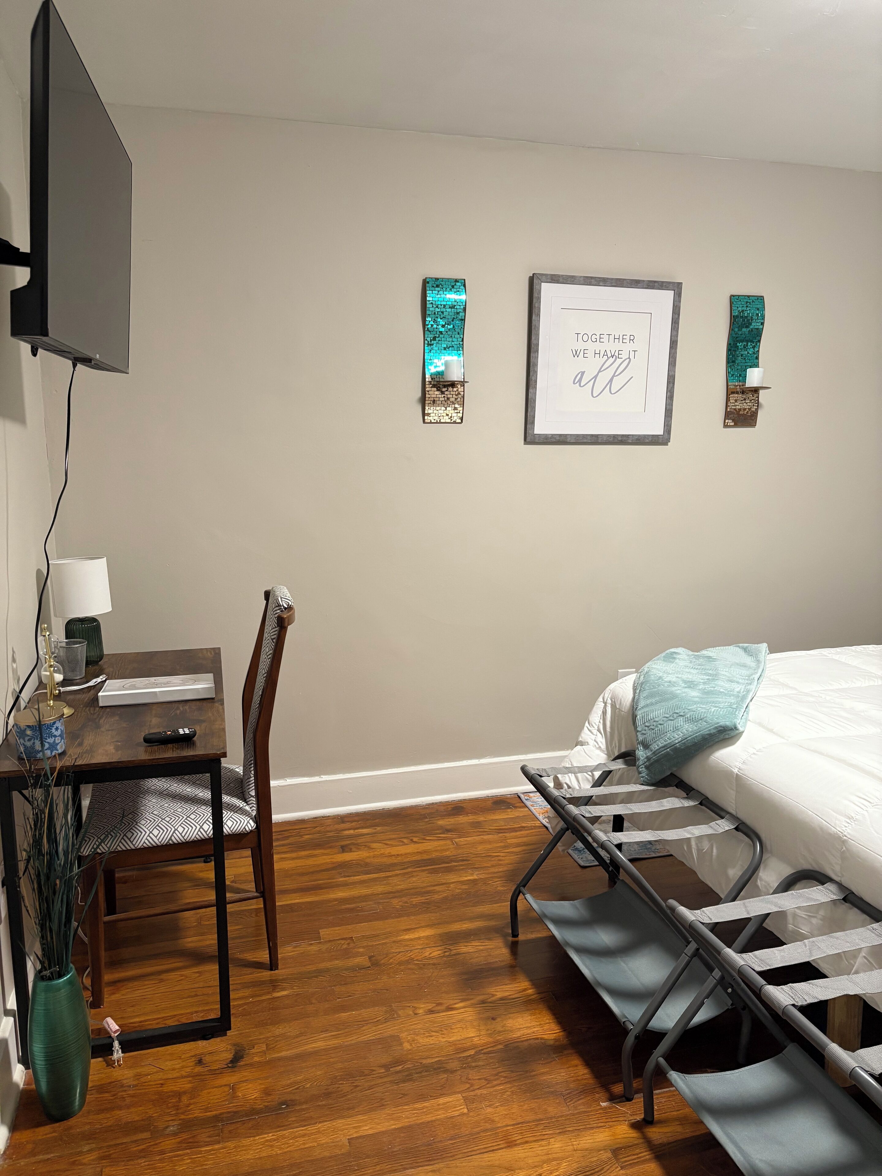 Una habitación con una cama, un escritorio y una silla. Hay un televisor montado en la pared, una cita enmarcada y un adorno en la pared.