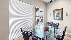Un coin repas avec une table et des chaises en verre, des stores blancs et un tableau encadré au mur.
