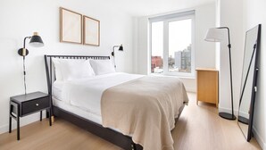 A modern bedroom with a large bed, bedside tables, a nightstand, a floor lamp, a desk, and a view of the city through a large window. - Blueground | Gowanus, view, nr Prospect Park (New York)