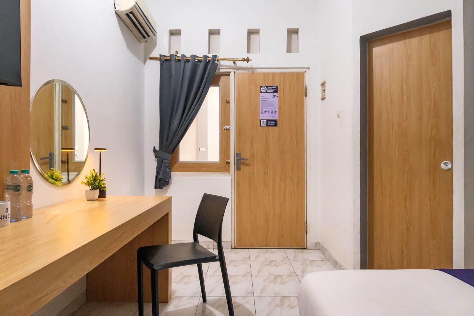 A hotel room with a wooden desk, a chair, a bed, a mirror, and a door with a sign.