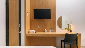 A hotel room with a wooden desk, a flat-screen TV, a round mirror, a chair, and a bed.