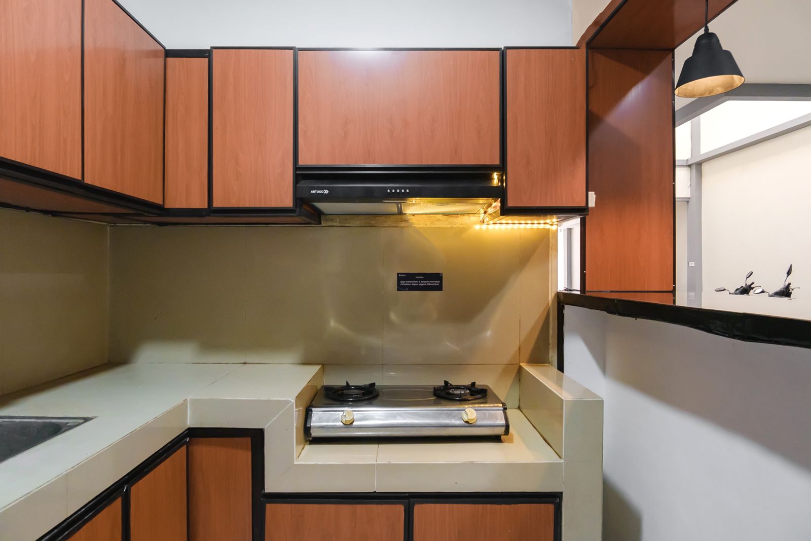 A compact kitchen with wooden cabinets, a stainless steel range, and a black pendant light.