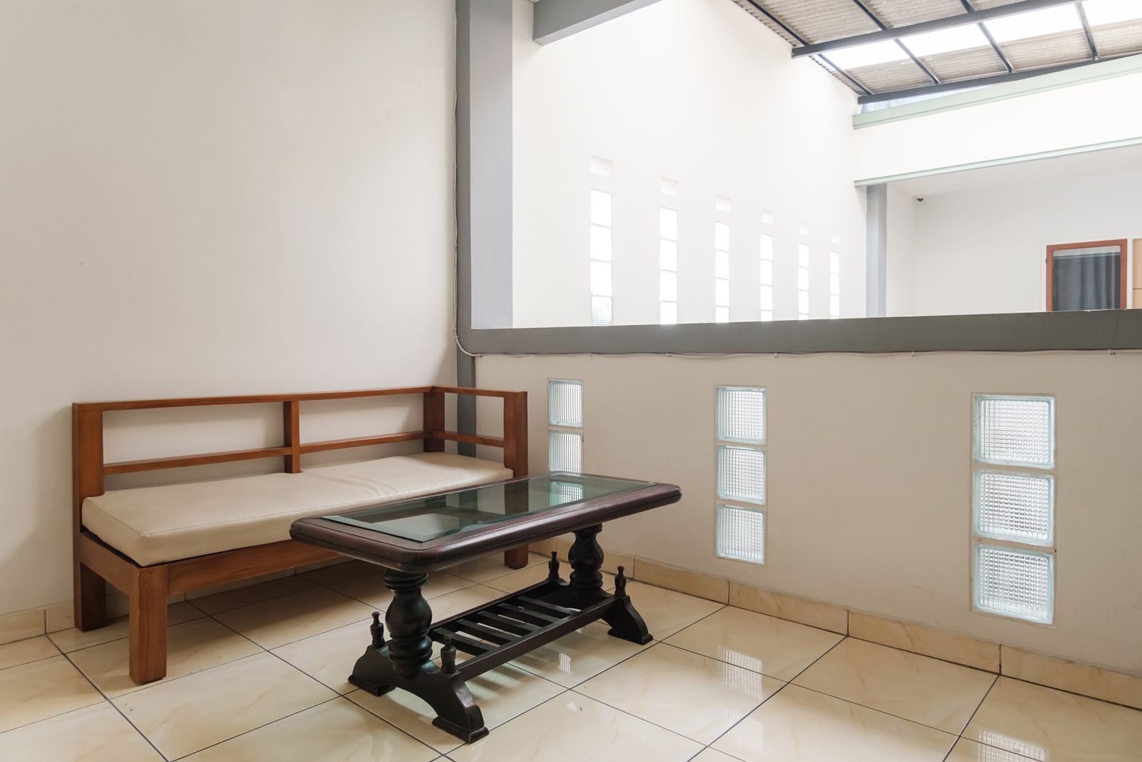 Una habitación con un banco de madera, una mesa con superficie de vidrio y vistas a un edificio a través de una ventana con persianas.