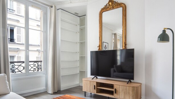 Un salon comprenant un grand téléviseur à écran plat posé sur un meuble en bois, un canapé, une table basse avec des magazines, un grand miroir et un balcon doté d’une balustrade.