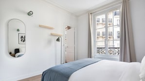Une chambre moderne avec un grand lit, un miroir, une table de chevet et une fenêtre donnant sur des immeubles.