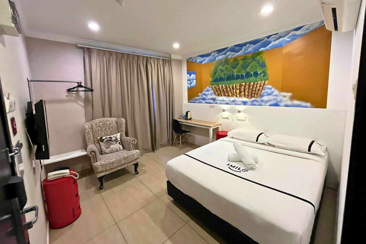 A hotel room with a bed, a desk, a chair, a red bucket, a television, and a wall mural.