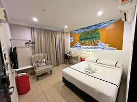 A hotel room with a bed, a desk, a chair, a red bucket, a television, and a wall mural.