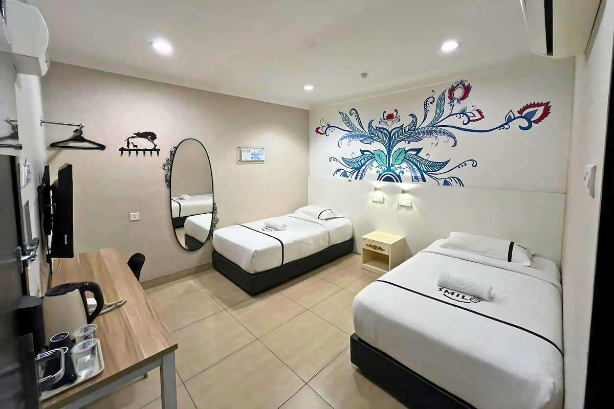 A hotel room with two beds, a desk with a kettle, a mirror, and a wall-mounted heater.
