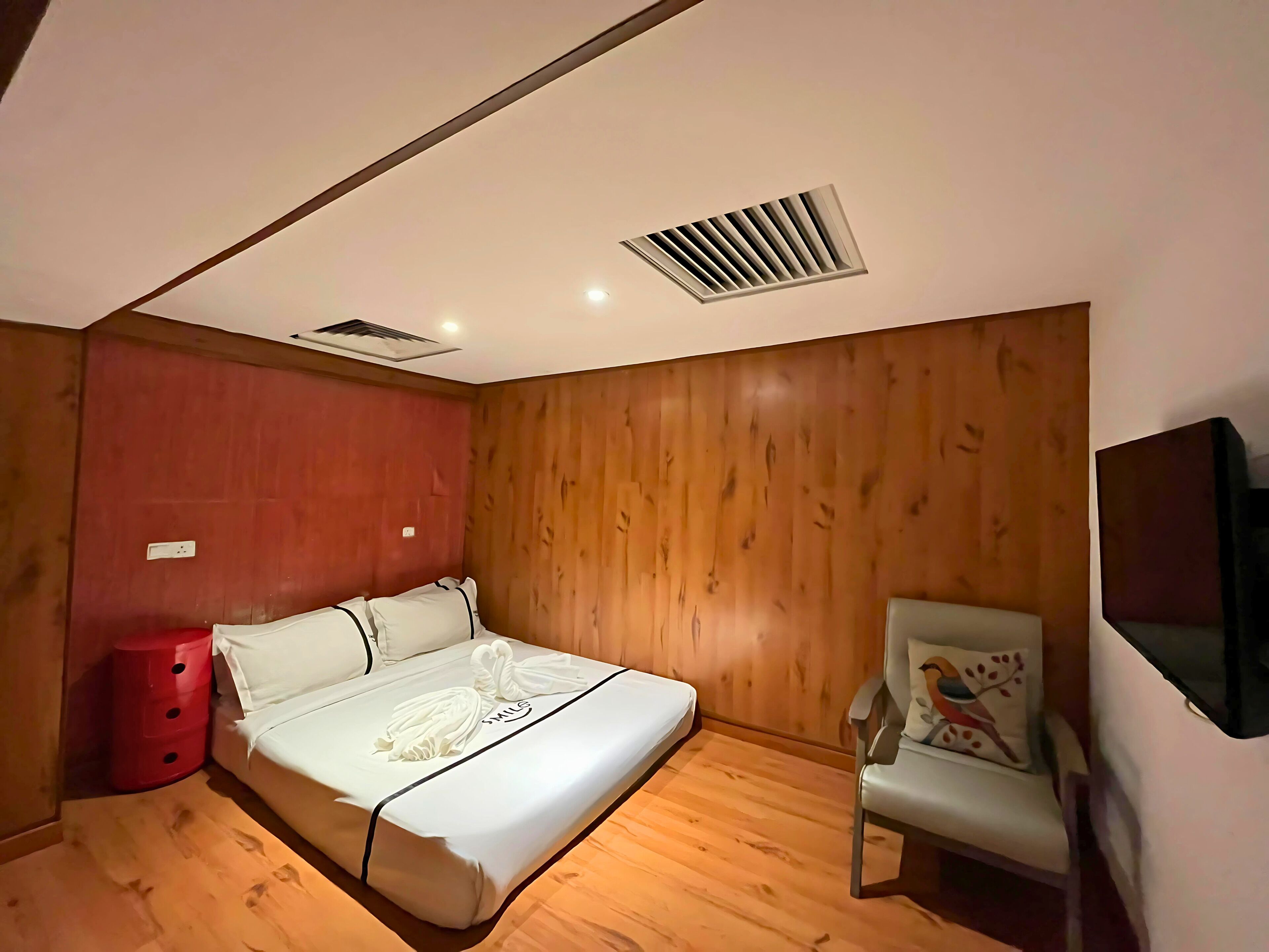 A bedroom with a bed, a red side table, a chair, and a TV mounted on the wall.