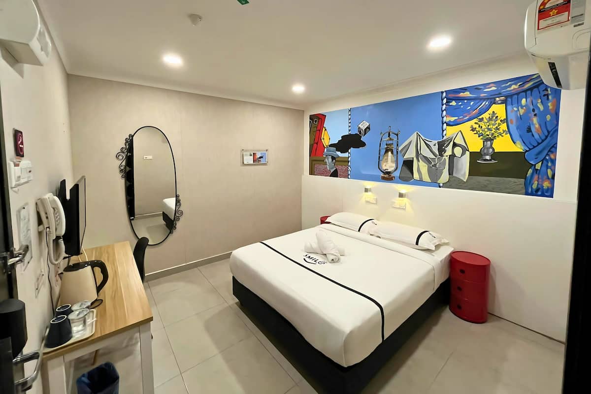 A hotel room with a bed, a desk with a telephone, a mirror, a wall-mounted air conditioner, and a colorful painting.