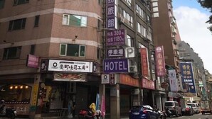 Un hôtel à plusieurs étages, dont la façade est ornée d’une grande enseigne indiquant « HÔTEL ». Chaque étage compte de nombreuses fenêtres et l’extérieur du bâtiment est en briques.
