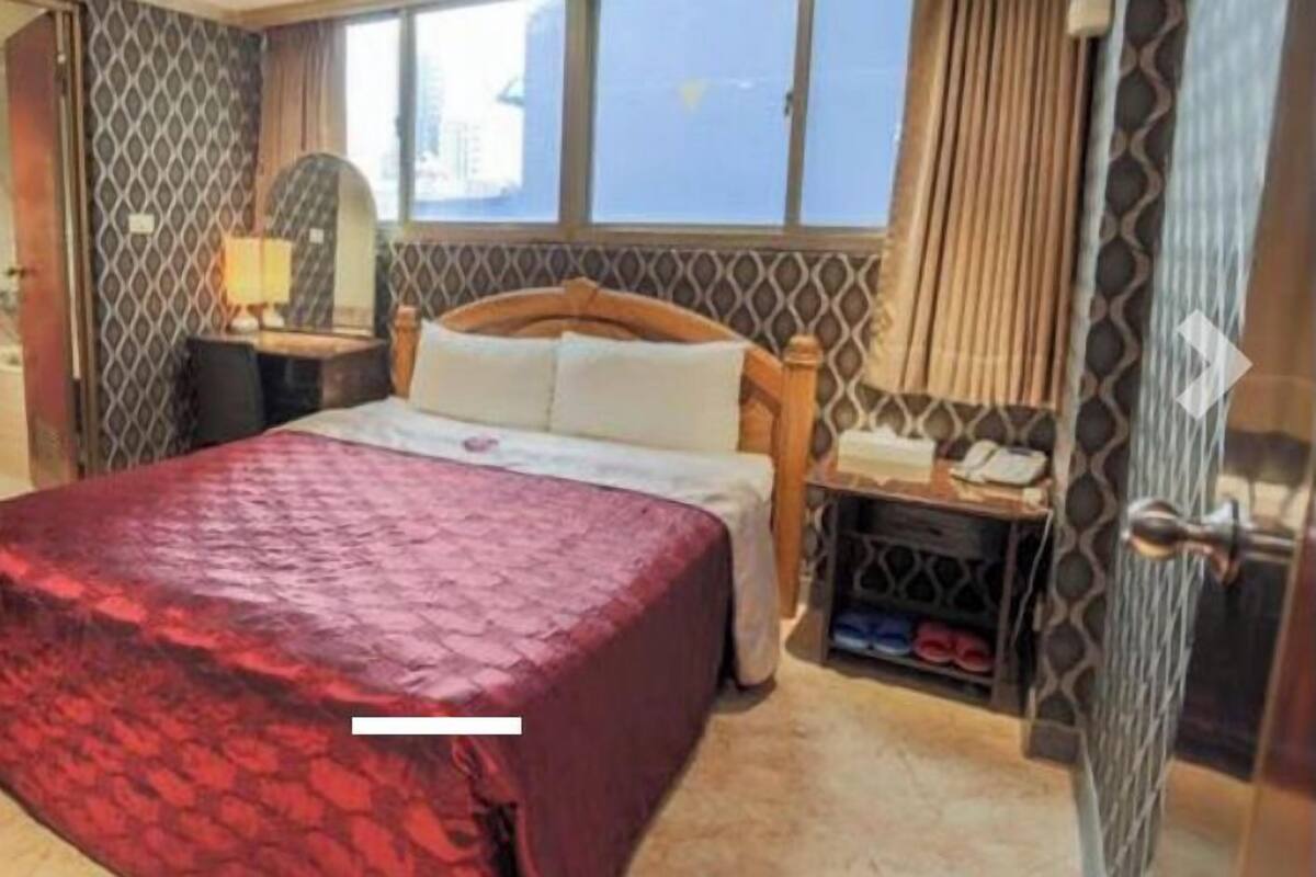 A hotel room with a bed, a nightstand, a mirror, a window, and a shoe rack.
