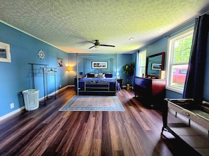 A bedroom with a four-poster bed, a ceiling fan, a window with curtains, a dresser, a mirror, and a plant.
