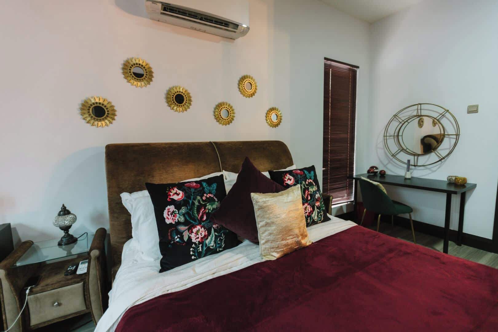 A bedroom with a large bed, a desk, and a chair. There are decorative wall mirrors and a wall-mounted air conditioner.