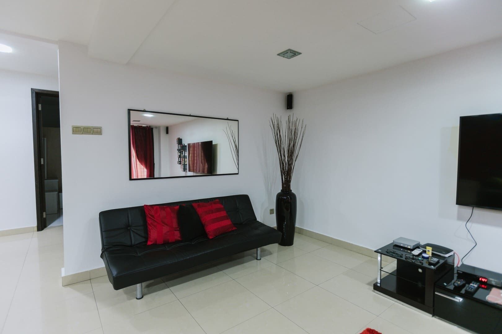 A modern living room with a black sofa, red cushions, a large mirror, a tall vase with branches, and a flat-screen TV.