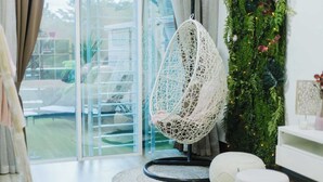 Una poltrona sospesa bianca in una stanza moderna con una porta vetrata che si apre su un giardino, un poggiapiedi bianco rotondo e una parete verde con piante.