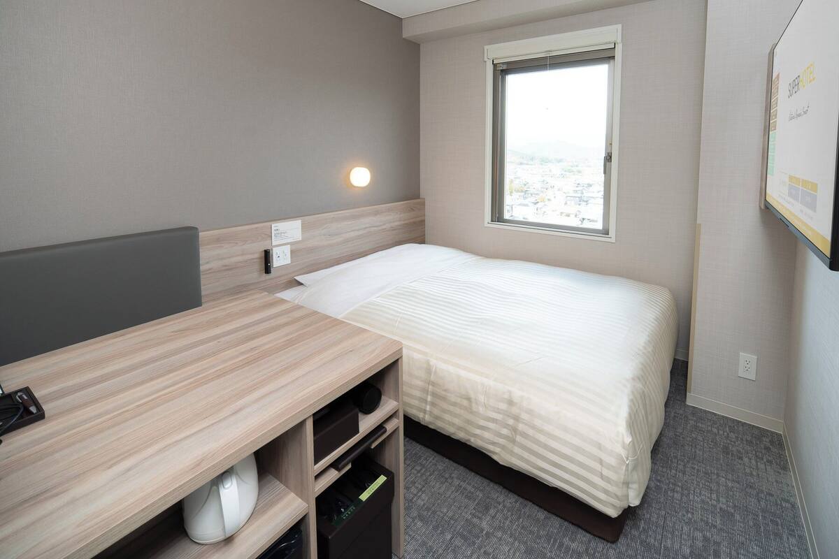 Habitación de hotel con cama, escritorio, silla, ventana con vistas y una pantalla en la pared.