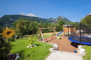 Ein Spielplatz mit Schaukeln, einer Rutsche und einem Trampolin, umgeben von einer Wiese und mit Bergblick.