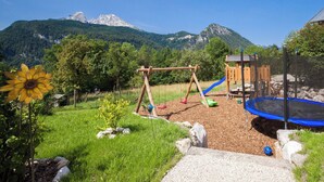 Un parque infantil con columpios, tobogán y trampolín, rodeado de césped y con una montaña al fondo.