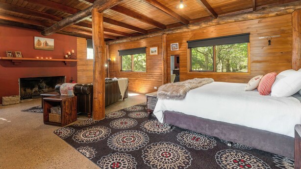 A log cabin bedroom with a fireplace, a large bed, and a view of trees through the windows.