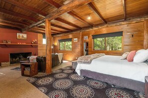 Un dormitorio tipo cabaña de madera con chimenea, una cama grande y vista a los árboles a través de las ventanas.