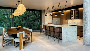 Una cocina moderna con una isla de mármol, iluminación colgante y vistas a abundante vegetación.