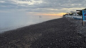 A pebbled beach with a calm sea, a cloudy sky, and a signboard on the right. - Qchen Cuan Cabin Amed  (Karangasem)