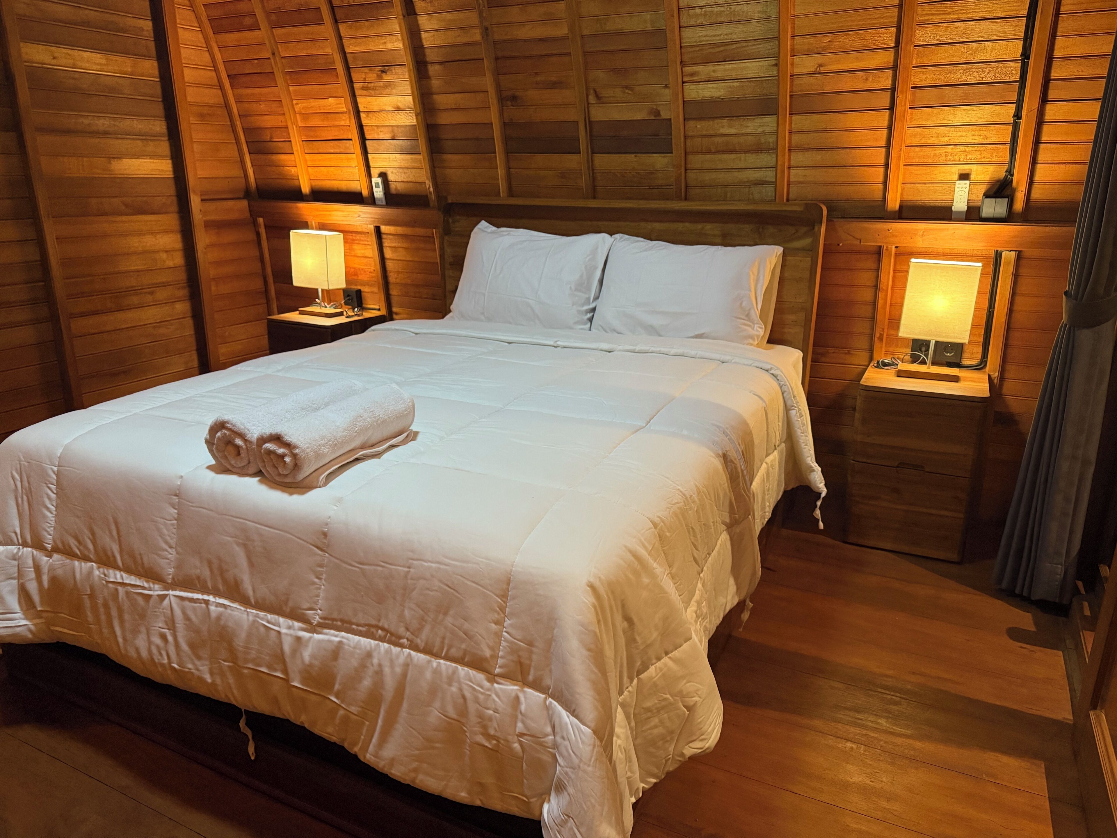 A neatly made bed with white linens and a rolled towel, wooden walls, and two bedside lamps.