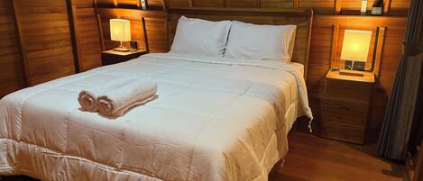 A neatly made bed with white linens and a rolled towel, wooden walls, and two bedside lamps.