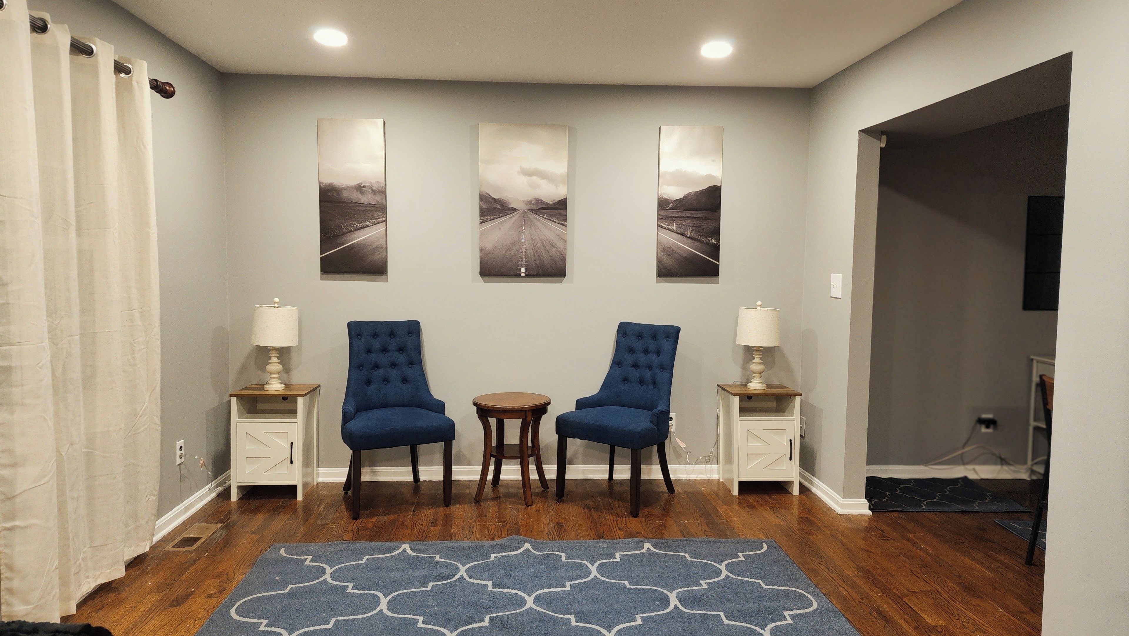 A room with two blue chairs, a small round table, and a patterned rug. There are three wall-mounted pictures depicting roads.