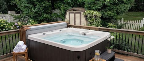 A hot tub on a wooden deck with a small table and towels, surrounded by greenery.