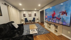 Una sala de estar con un sofá negro, una mesa de centro, un televisor de pantalla plana y dos sillones.