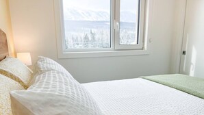A bed with white bedding and a view of snow-covered mountains through a window. - Casa del Oso - DT Condo, River & Mountain Views (Golden)