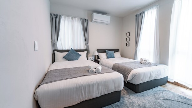 A modern hotel room with two beds, a large window with curtains, an air conditioning unit, and a small bedside table with a lamp. - GUEL PALACIO (Miyakojima)